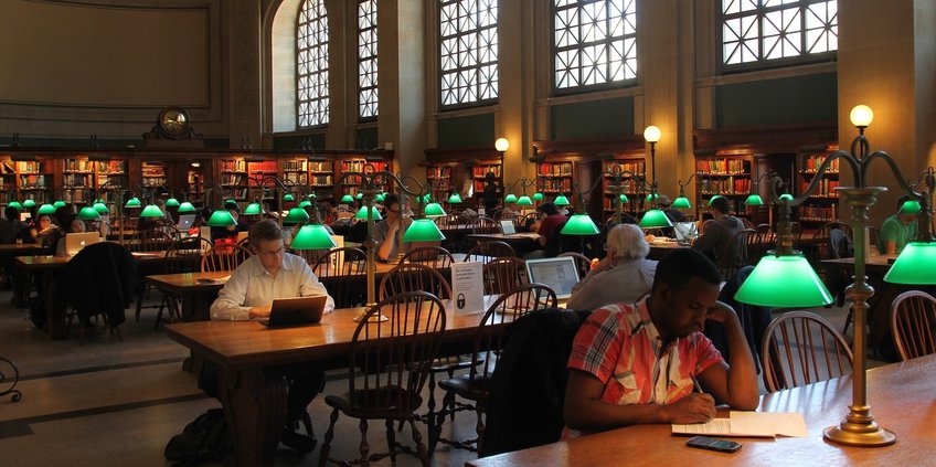 A view in a large, traditional library reading room with high windows and roof.
In the middle there a rows of long tables of wood with green lamps.There are some user´sitting at the tables, working with their laptop or writing.