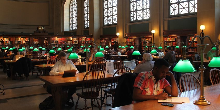 Find the information you are looking for! A view in a large, traditional library reading room with high windows and roof.
In the middle there a rows of long tables of wood with green lamps.There are some user´sitting at the tables, working with their laptop or writing.