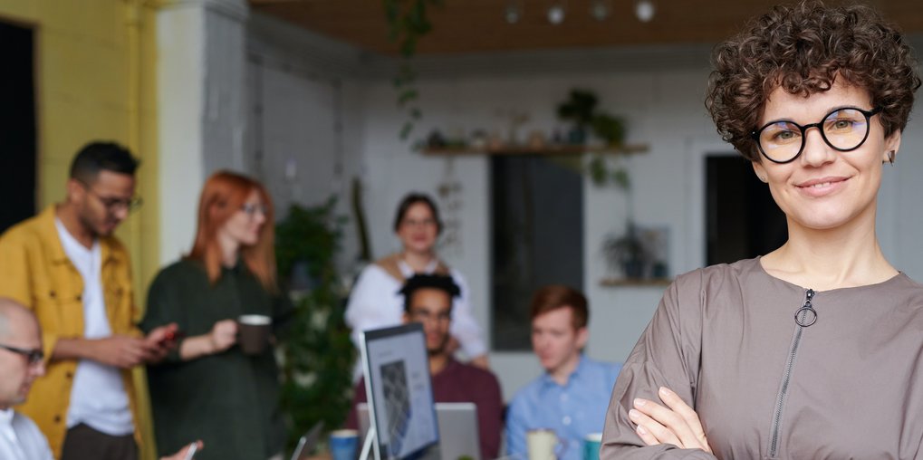Rechts im Vordergrund ist eine junge, lächelnde Frau mit Brille und kurzem braunem, gelocktem Haar abgebildet. Im Hintergrund verschwommen ist eine Gruppe von stehenden und sitzenden jungen Menschen in einem Büro dargestellt.