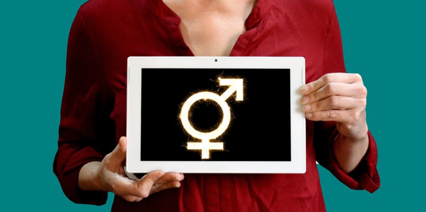 Ein Foto von einer schwarzen Tafel mit einem weißen Symbol, das sowohl für das weibliche als auch das männliche Geschlecht steht. Die Tafel wird von einer Person gehalten, von der man nur den Oberkörper im Hintergrund sieht.