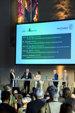 Eine Podiumsdiskussion mit fünf Rednern auf einer Bühne, die vor einem Publikum sprechen. Im Hintergrund ein beleuchtetes Kirchenfenster. Ein Bildschirm zeigt Namen und Titel der Beteiligten an, darunter Dietmar Harhoff, Direktor am Max-Planck-Institut für Innovation und Wettbewerb, Susanne Kadner, Co-Founder CIRCULAR REPUBLIC & Directorin der UnternehmerTUM, Kai Sundmacher, Direktor am Max-Planck-Institut für Dynamik komplexer technischer Systeme und Ralf P. Thomas, Finanzvorstand der Siemens AG