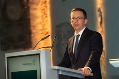 A man in a dark suit stands at a lectern bearing the Max Planck logo, with microphones and an illuminated background.